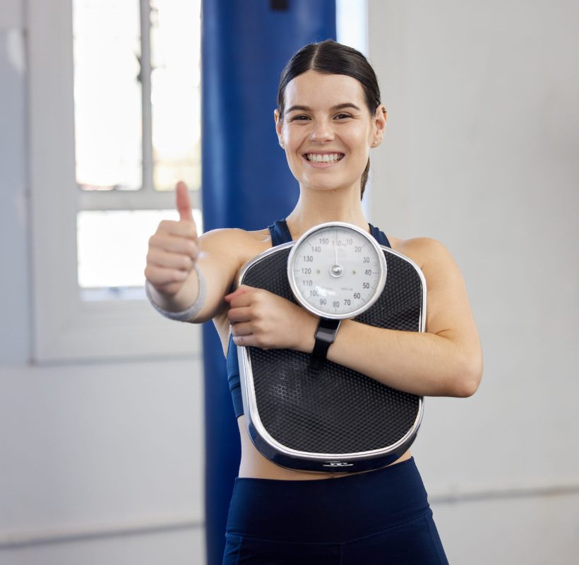 Woman, gym portrait and thumbs up with scale, wellness and weight loss goal for health, body or fit.